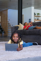 Naklejka premium African American children lying on plush rug in living room, holding tablet and smartphone