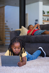 Naklejka premium Teenage girl lying on rug using tablet, sibling reclining on sofa with smartphone in living room
