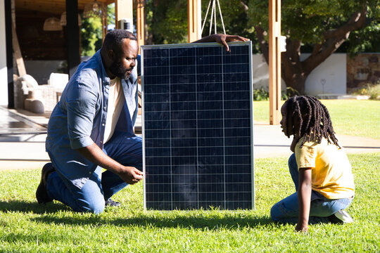 African American father and son kneeling and holding solar panel on backyard lawn near porch