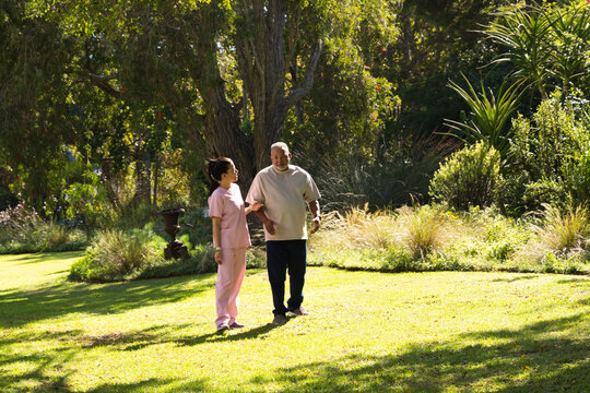 Asian caregiver wearing pink scrubs guiding African American senior on sunlit lawn by tree and urn