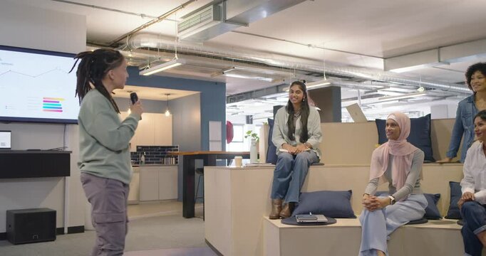 Nonbinary African presenter opening talk, holding mic, speaking to seated team in office