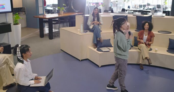 Diverse coworkers clapping at office, nonbinary presenter rising, walking center, speaking into mic