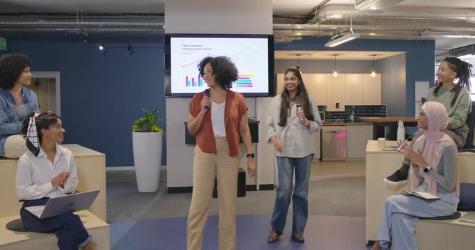 Colleagues clapping in open-plan lounge, sending two women presenting charts with microphones