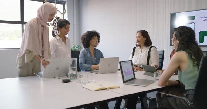 Diverse female coworkers entering meeting room, two women placing laptops, starting team discussion