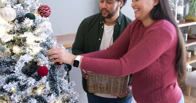 African American couple decking tree at home, woman in maroon sweater hanging ornaments from basket