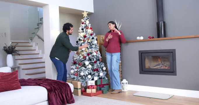 African American couple decorating Xmas tree at home, woman handing red bauble, man adding baubles