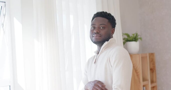 African Adult male pausing by large window daylight drawing eye, turning to cam, smiling in sweater