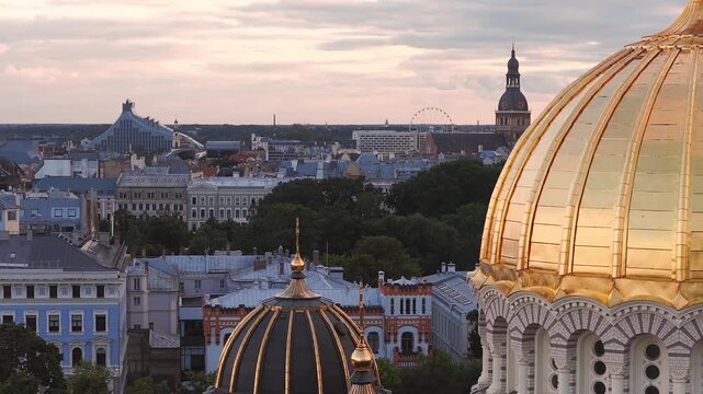 An aerial panning shot captures the golden dome of Riga Nativity Cathedral, historic rooftops, and the National Library in Latvia.