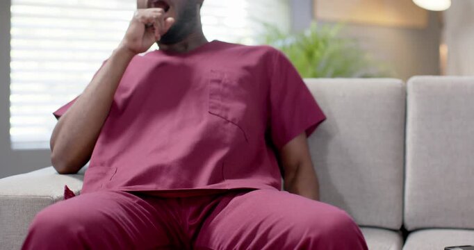 African American man in maroon scrubs fidgeting with tablet on sofa, yawning and dozing after shift