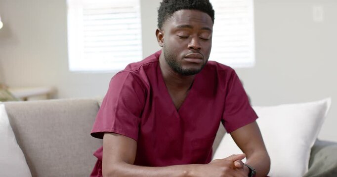 African American man in maroon scrubs sitting on sofa at home, blinking, reaching, retrieving item