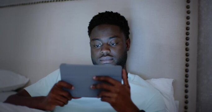 African American adult man reclining on bed holding tablet, blue screen causing scrolling and smile