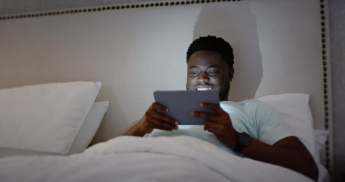 African American man reclining in bed holding tablet, watching funny clip, laughing and tapping