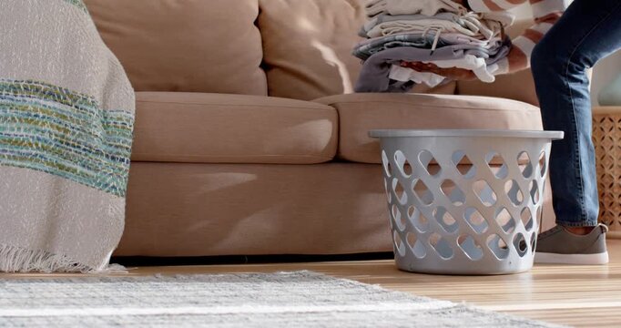 Person appearing from right is lifting folded laundry on couch and placing into basket to organize