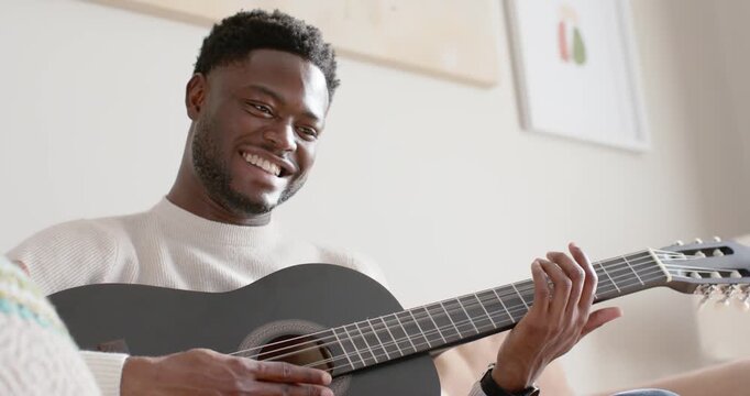 African American man 20s laughing, strumming black guitar at home practicing song in sweater, watch