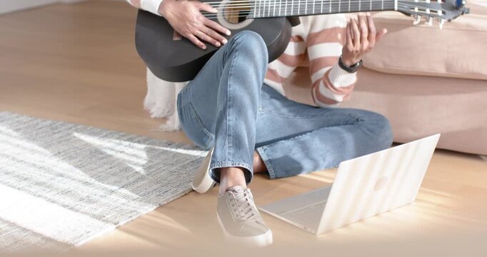 Woman is tapping laptop to start lesson on hardwood floor while forming chords and strumming guitar
