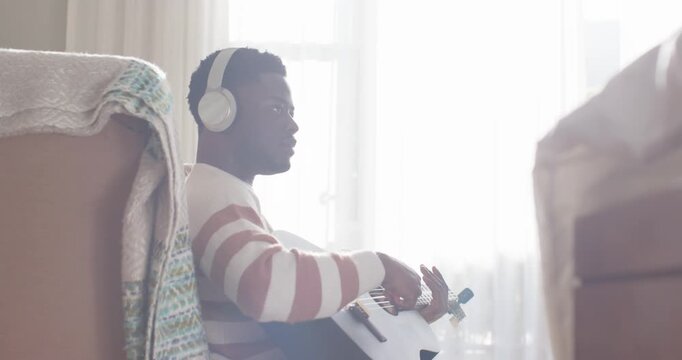 African American man settling with headphones, adjusting guitar, strumming, practicing near window