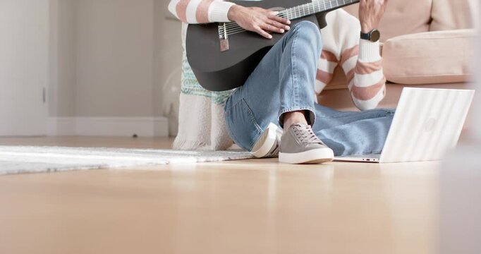 Seated musician is referencing laptop and strumming black guitar on floor, practicing chord changes