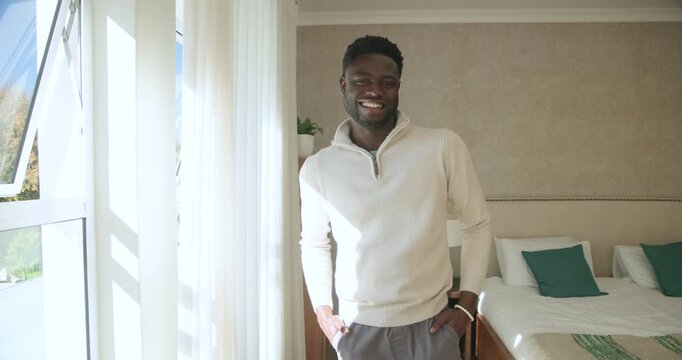 African American man in his 20s noticing camera, stepping in bedroom near window and bed, smiling
