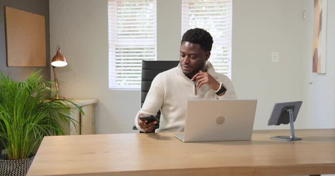 African American man in pullover working at laptop hearing phone buzzing, answering call by blinds