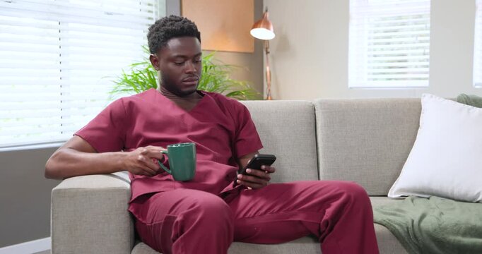 African American man sitting gray sofa in burgundy scrubs, smiling reading phone, holding green mug