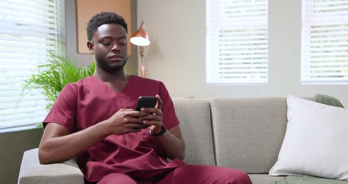 African American man in maroon scrubs scrolling phone on gray sofa during break, drifting to sleep