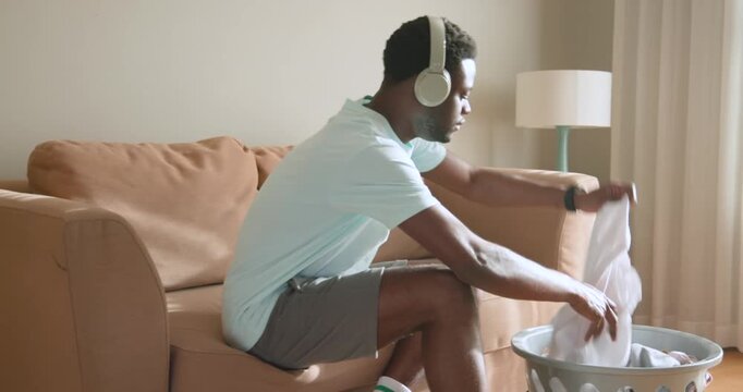 African American adult male carrying basket, sitting in living room folding shirts, sorting clothes