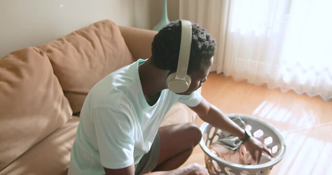 African American man sitting on sofa with headphones reaching into basket folding shirt to organize