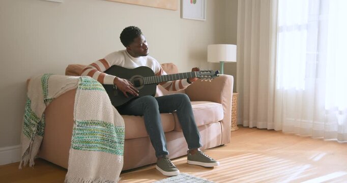 African man sitting upright on pink sofa at home practicing on black guitar, tuning, checking frets