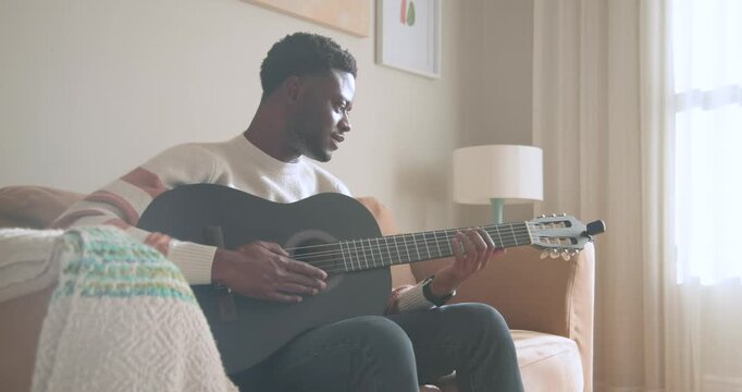 African American man in cream sweater smiling, practicing chords on black guitar on sofa by window