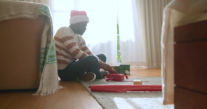 African American man in Santa-hat sitting by window on rug, tying ribbon on gift for Christmas