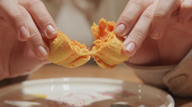 Close up of hands holding and breaking yellow macaron revealing its soft texture and orange filling. scene features natural light highlighting detailed interaction between fingers and food