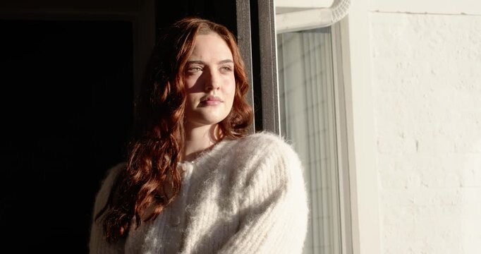 Woman leaning in jamb by window, sun streaming, opening eyes, turning toward light in fuzzy sweater