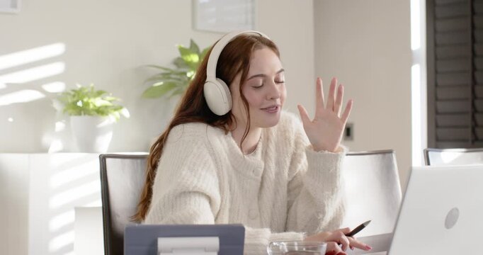 Woman in 20s at home wearing headset, joining online meeting, gesturing with pen near laptop
