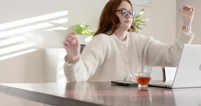 Woman in knit sweater after stretching, typing on laptop at wood table teacup phone visible
