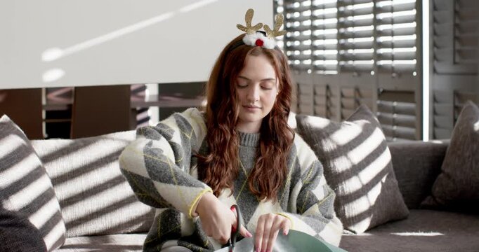 20s woman with reindeer headband on sofa cutting wrapped gift to open with scissors under sunlight