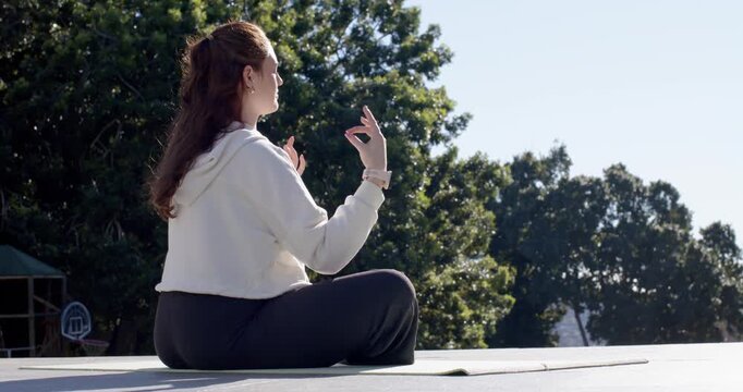 Female starting meditation on mat on deck in white hoodie and dark leggings, forming mudras