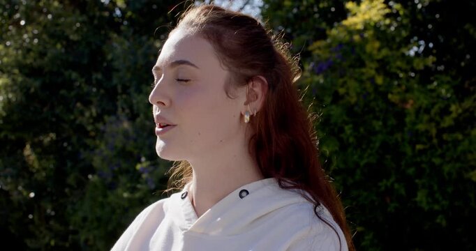 Woman standing in garden, pausing then breathing while turning to profile, sunlight on hoodie hoops