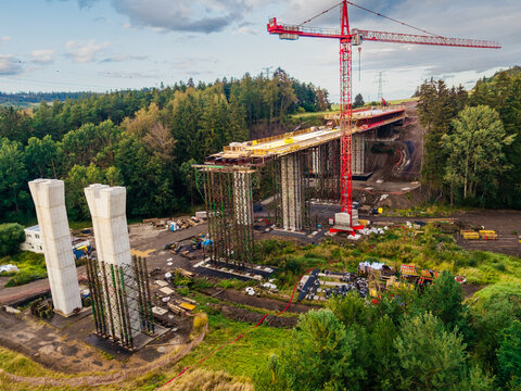 Aerial view of the Lezky bridge construction site featuring a tall red crane, concrete bridge piers, and scaffolding, surrounded by lush green forests in Lubenec, Usti nad Labem Region, Czechia.