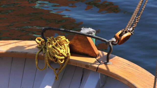 Close-up of a classic wooden sailboat's bow with ropes and pulley, gently rocking on calm water with reflections.