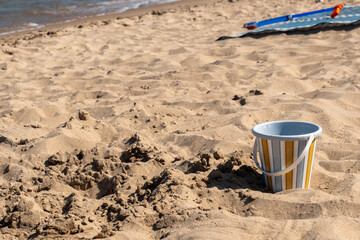 Colorful striped bucket on a sunny sandy beach, representing summer activities, beach toys, family vacation, seaside leisure, outdoor recreation and fun beach activities © Juli