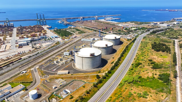 Aerial view of large oil storage tanks at the industrial petroleum terminal, with shipping port cranes and containers visible in the background in Sines, Setubal, Portugal.
