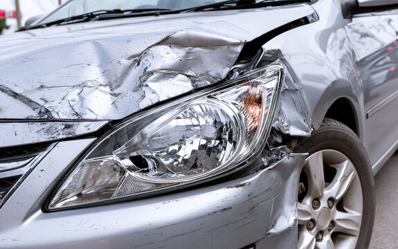 Close up view of damaged silver car after accident. Crumpled metal hood, smashed fender show heavy impact. Broken headlight, dented bumper near wheel at roadside. Vehicle needs expensive repair due