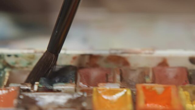 Close up of paintbrush mixing colors in watercolor palette with various hues. brush repeatedly dips into different color wells demonstrating process of selecting and combining paints