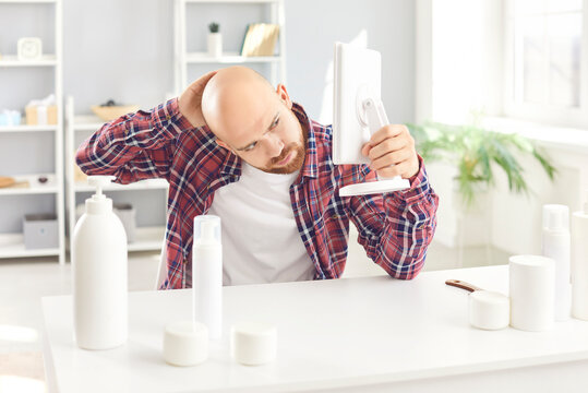 Young bearded man wearing casual clothes with bald head looking in the mirror sitting at table with cosmetics bottles at home. Male person preparing for beauty spa day. Skin and body care concept