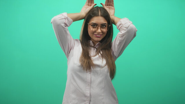 Young woman in glasses raises hands behind head making rabbit ears gesture against a mint green backdrop in studio; playful energy.