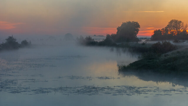 A serene river is enveloped in thick morning mist during a vibrant orange sunset, with dark silhouettes of distant trees