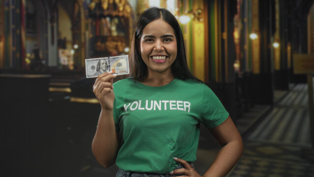 Young volunteer woman holding a hundred dollar bill with hand to mouth surprise gesture in a museum gallery with ornate columns and patterned floor; surprise generosity.