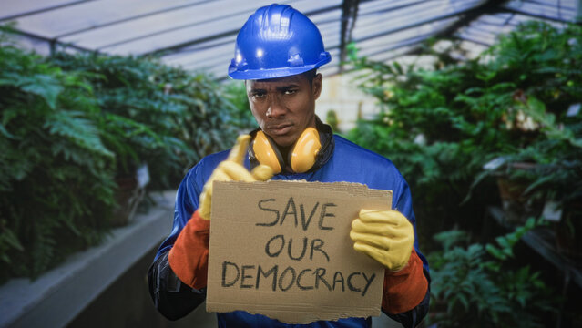 Man construction worker holding cardboard sign and pointing finger in greenhouse; solidarity civic duty.