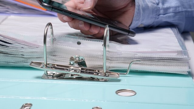 Businessperson's hand holding a smartphone to scan a document lying on top of a ring binder, integrating digital and physical record keeping for modern office work