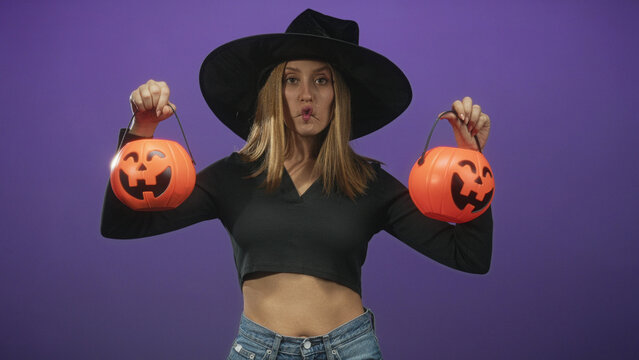 Woman in witch hat holds two jack o lantern buckets by handles, puckered lips and exposed midriff against purple studio wall; playful mischief.